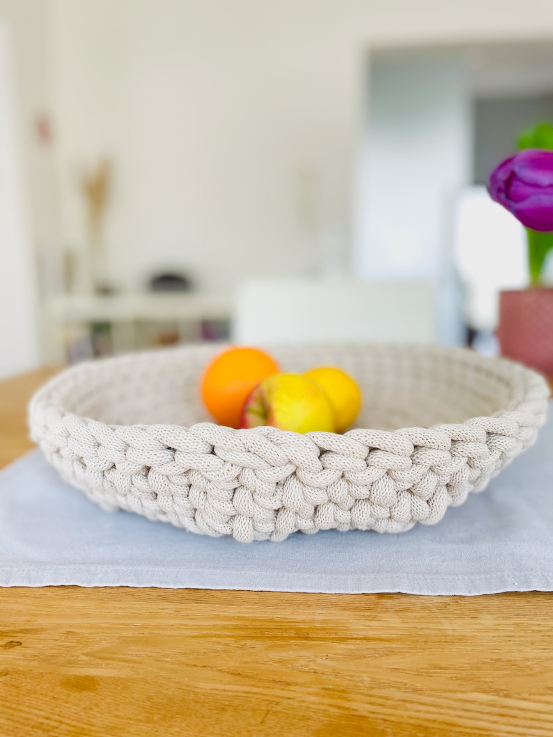 K&ouml;nnte beinhalten: Eine beige geh&auml;kelte Schale gef&uuml;llt mit Orangen und &Auml;pfeln steht auf einem hellblauen Platzdeckchen auf einem Holztisch.