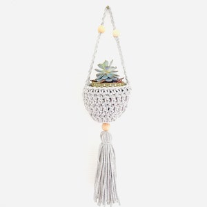 Puede incluir: Un macetero de macramé gris con un acento de cuentas de madera y un borla. Una pequeña planta suculenta está en la maceta.