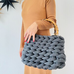 May include: A gray knitted tote bag with a bamboo handle. The bag is made of thick, chunky yarn and has a woven texture.