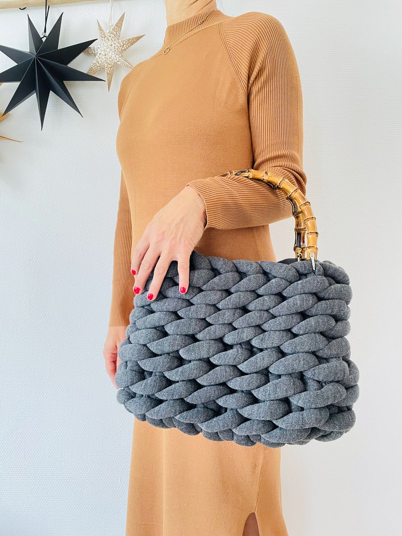 May include: A gray knitted tote bag with a bamboo handle. The bag is made of thick, chunky yarn and has a woven texture.