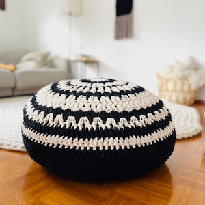 May include: A black and white crocheted round pouf or ottoman with a geometric pattern. The pouf is sitting on a wooden floor.