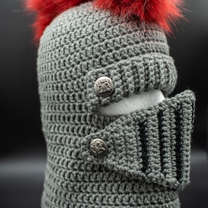 Puede incluir: Un casco de caballero de ganchillo gris con una pluma de piel sintética roja. El casco presenta una visera con rayas negras y se sujeta con dos botones decorativos. El artículo está hecho a mano.