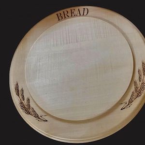 May include: A round wooden bread board with a raised centre. The word "BREAD" is etched in the wood along the top edge. Wheat stalks are etched on the sides. The board is light brown.