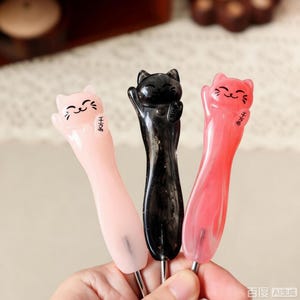 May include: Three crochet hooks with handles shaped like smiling cats. The handles are light pink, black with subtle glitter, and bright pink. Each cat figure has small ears, whiskers, and a smiling face with Japanese characters on the side.
