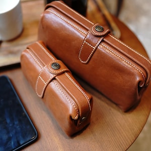 May include: Two brown leather cases of different sizes, featuring white stitching and a clasp closure with a metal button. The cases are on a wooden surface, suggesting a handcrafted or vintage aesthetic. The leather has a rich, warm tone.