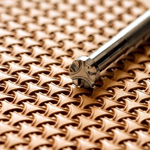 May include: A close-up of a silver leather stamping tool with a cross-shaped design. The tool is resting on a piece of brown leather that has been embossed with a repeating pattern.