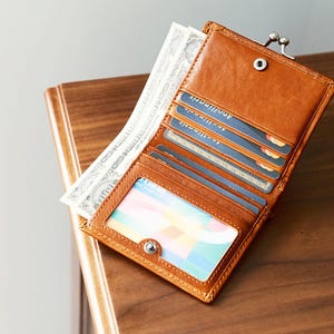 Könnte beinhalten: Geöffnetes, braunes Lederportemonnaie mit mehreren Kartenfächern und einem transparenten Ausweisfach. Das Portemonnaie ist mit Kreditkarten und Bargeld gefüllt. Es hat einen Metallverschluss und liegt auf einer Holzoberfläche.