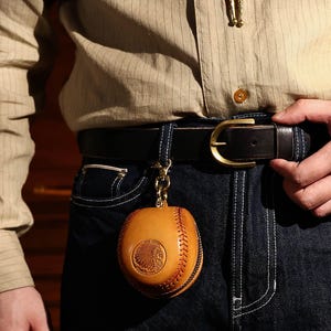 May include: A brown leather baseball-shaped keychain with a stitched design and an embossed Native American head logo. The keychain is attached to a black leather belt with a gold buckle.