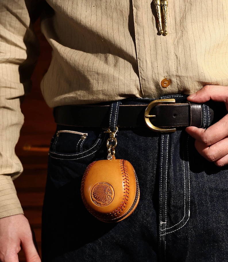 May include: A brown leather baseball-shaped keychain with a stitched design and an embossed Native American head logo. The keychain is attached to a black leather belt with a gold buckle.