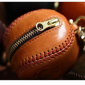 May include: A brown leather baseball-shaped zippered pouch with red stitching. The zipper is open and the pouch is empty.