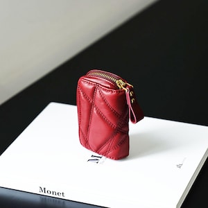 May include: A small, quilted, claret leather pouch with a gold zip and a small strap. The pouch is rectangular with a diamond pattern and is on a white surface with the word "Monet" printed on it.