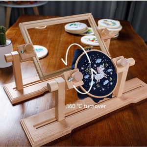 May include: A wooden embroidery hoop stand with a rotating mechanism. The stand is made of natural wood and has a 360 degree turnover feature. The stand is holding a hoop with a blue fabric and a unicorn embroidery design.