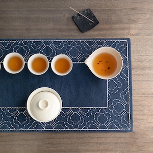May include: A tea set arranged on a navy blue mat with a white embroidered border. The set includes small white teacups filled with amber tea, a teapot with a lid, and a serving bowl. An incense holder and incense stick are also present.