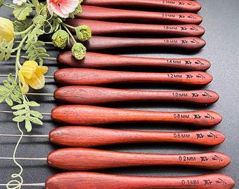 Op de afbeelding: Een set van elf haaknaalden met donker roodbruine houten handvatten, elk voorzien van een metalen punt en maataanduidingen variërend van 0,1 mm tot 2,8 mm. Een decoratieve gehaakte bloementak met roze, gele en groene garenaccenten is bevestigd.