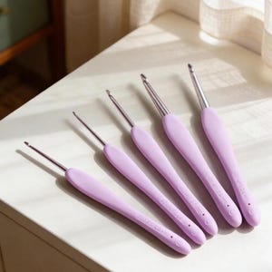 Peut inclure: Un ensemble de cinq crochets avec des pointes en métal argenté et des manches ergonomiques violet clair. Chaque manche présente un texte en relief indiquant la taille en millimètres, de 2,0 mm à 6,0 mm, avec une petite encoche circulaire.