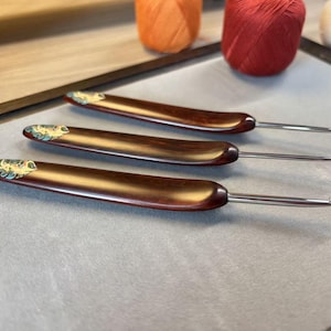 Puede incluir: Tres ganchillos con mangos de madera marrón oscuro y puntas de metal plateado. Cada mango presenta una incrustación decorativa en el extremo con un patrón colorido y arremolinado en tonos azul, verde y naranja sobre un fondo claro.