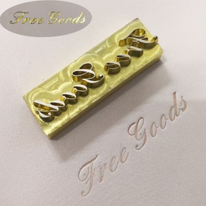 Op de afbeelding: Een goudkleurige metalen stempel met de woorden "Free Goods" in een cursief lettertype. De stempel is rechthoekig met verhoogde letters in een gestileerd lettertype. De gestempelde afdruk van de woorden is eronder zichtbaar.