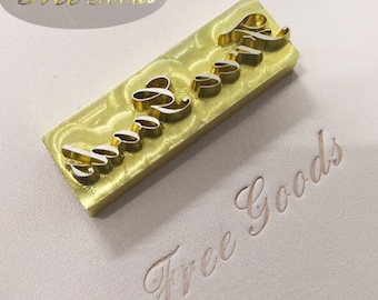 May include: A gold-colored metal stamp with the words "Free Goods" in a cursive font. The stamp is rectangular with the raised letters in a stylized font. The stamped impression of the words is visible below the stamp.