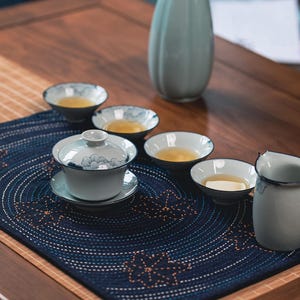 May include: A tea set on a wooden table. The set includes a blue and white tea cup with a lid, small bowls, a pitcher, and a tall vase. The tea is a golden colour. The set is on a navy blue placemat with a floral design.