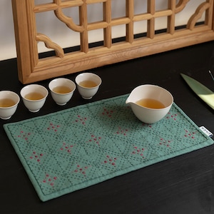 May include: A tea set arrangement featuring small white teacups and a serving bowl filled with tea. A green, patterned placemat with red floral accents sits beneath the tea set. A wooden window frame is in the background.