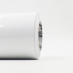 Peut inclure: Un rouleau de film adhésif en vinyle blanc. Le film est enroulé sur un noyau en carton avec du texte noir.
