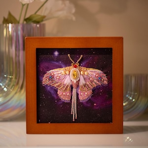 Könnte beinhalten: Gerahmtes Kunstwerk mit einem bestickten Motten-Design. Die Motte hat rosa und goldene Flügel mit detaillierter Perlenarbeit, vor einem lila Galaxiehintergrund. Der Holzrahmen verleiht diesem dekorativen Stück einen klassischen Look.