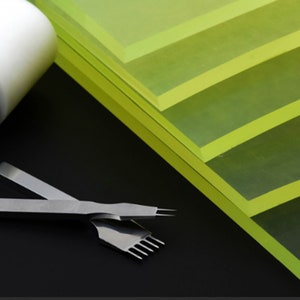 May include: A set of four green acrylic cutting mats stacked on top of each other. A leather working tool with a wooden handle and a white head is lying on the left side of the image. A leather working tool with a metal handle and a metal blade is lying on the right side of the image.