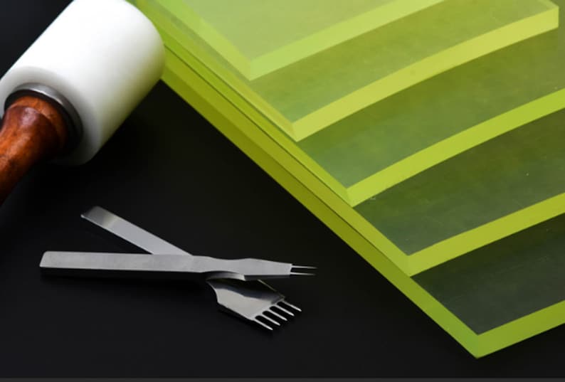 May include: A set of four green acrylic cutting mats stacked on top of each other. A leather working tool with a wooden handle and a white head is lying on the left side of the image. A leather working tool with a metal handle and a metal blade is lying on the right side of the image.