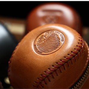 May include: A brown leather baseball-shaped coin purse with a red stitching detail and a zipper closure. The purse features an embossed design of a Native American chief's head with the text "Red Moon Co. Est. 2015".