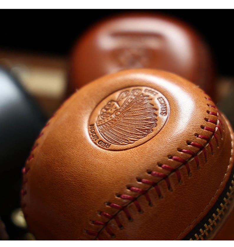 May include: A brown leather baseball-shaped coin purse with a red stitching detail and a zipper closure. The purse features an embossed design of a Native American chief's head with the text "Red Moon Co. Est. 2015".