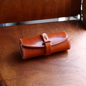 Può includere: Un astuccio arrotolabile in pelle marrone con una fibbia in ottone e cuciture bianche. L'astuccio è chiuso e si trova su una superficie in legno.