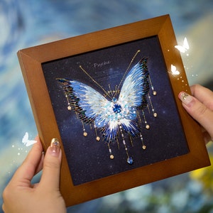 Pode incluir: Uma obra de arte emoldurada apresentando uma borboleta azul e branca com asas de contas e um corpo incrustado de cristais. A borboleta é representada sobre um fundo azul escuro com um padrão estrelado. A moldura é de madeira e tem um acabamento marrom. A obra de arte é intitulada "Psyche".