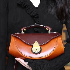 May include: A brown leather satchel handbag with a top flap closure and a brass lock. The bag has a curved shape and a short, sturdy handle.