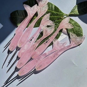 May include: A collection of crochet hooks with translucent, marbled pink handles shaped like mermaid tails. Each handle features a metallic hook tip and is imprinted with the text '金誉 2.0'. The hooks are arranged in a fan-like pattern.