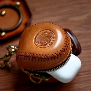May include: A brown leather case with a red stitching detail and a zipper opening. The case has a red logo with an image of a Native American headdress and the text "Red Moon Est. 1993 Since".
