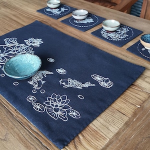 以下が含まれることがあります： 白い刺繍の鯉、蓮の花、葉が施された濃い青色のテーブルランナー。ランナーは木製のテーブルの上にあり、4つの小さなマッチングコースターがあります。