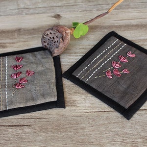 Puede incluir: Dos posavasos de lino gris con ribete negro y flores bordadas de color rosa.