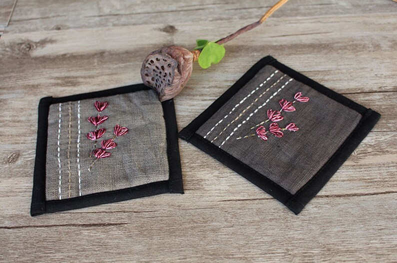 Puede incluir: Dos posavasos de lino gris con ribete negro y flores bordadas de color rosa.