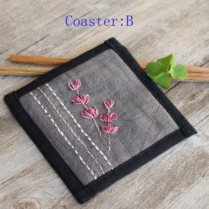 Puede incluir: Un posavasos de lino gris con un borde negro y flores rosas bordadas. El posavasos est&aacute; sobre una superficie de madera.