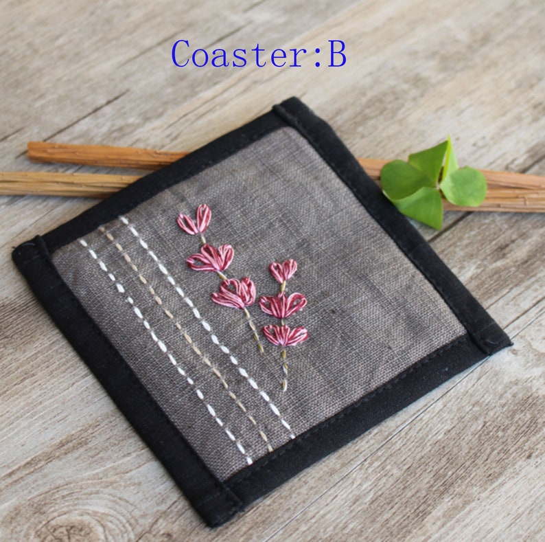 Puede incluir: Un posavasos de lino gris con un borde negro y flores rosas bordadas. El posavasos est&aacute; sobre una superficie de madera.