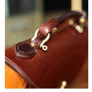 May include: Close-up of a brown leather handbag with a gold metal clasp and handle. The handle is attached to the bag with a decorative metal loop.