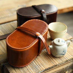 May include: Two leather boxes, one brown and one tan, with stitched edges and leather straps. A small white teapot with blue floral designs and two small cream-coloured teacups sit nearby, suggesting a tea set.