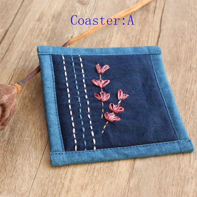 Puede incluir: Un posavasos de tela azul con un borde cosido y un peque&ntilde;o dise&ntilde;o de flor bordada en rosa.