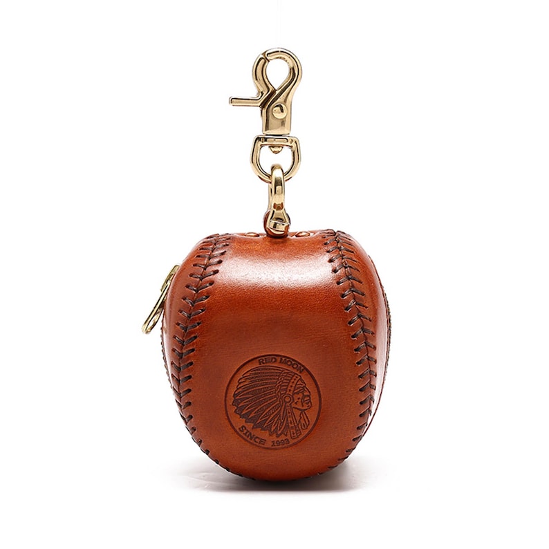 May include: A brown leather baseball-shaped keychain with a gold zipper and a gold clasp. The keychain has a stitched baseball seam design and a Red Moon logo with an American Indian head and the text "Since 1953".