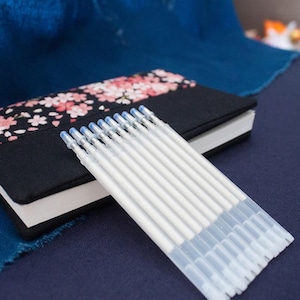May include: A pack of ten clear, white, refillable ink pens with blue tips. The pens are stacked on top of each other and are lying on a blue surface.