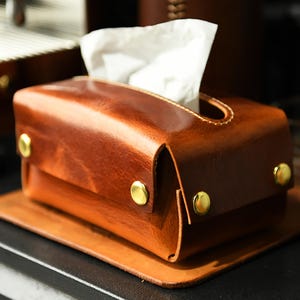 Puede incluir: Un dispensador de pañuelos de cuero marrón con forma rectangular y remaches dorados. El dispensador contiene pañuelos blancos y se apoya sobre una base de cuero a juego. El cuero tiene un tono cálido y rico.