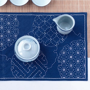 May include: A blue felt tea mat with white embroidered designs. The mat features three circular designs with a repeating flower pattern. A white ceramic teapot and a white ceramic teacup are placed on the mat.