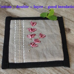 Puede incluir: Un posavasos cuadrado de lino gris con borde negro. El posavasos tiene un dise&ntilde;o bordado sencillo de un tallo de flor rosa con hojas.
