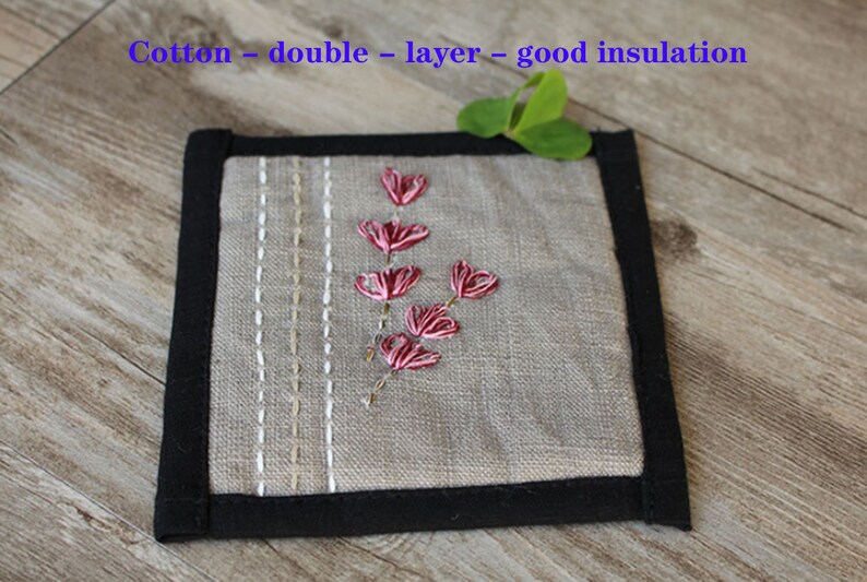 Puede incluir: Un posavasos cuadrado de lino gris con borde negro. El posavasos tiene un dise&ntilde;o bordado sencillo de un tallo de flor rosa con hojas.
