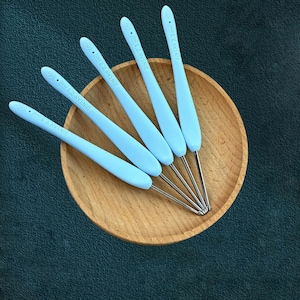 May include: Five light blue crochet hooks with silver metal tips are arranged in a wooden bowl. The hooks have the text "JIN LAN 2.3 mm" printed on them. The bowl sits on a dark green surface.